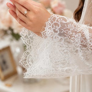 May include: A white lace robe featuring long, wide sleeves with intricate floral patterns and scalloped, fringed edges. The fabric is sheer and delicate, showcasing a detailed lace texture throughout the sleeve design and cuff area.