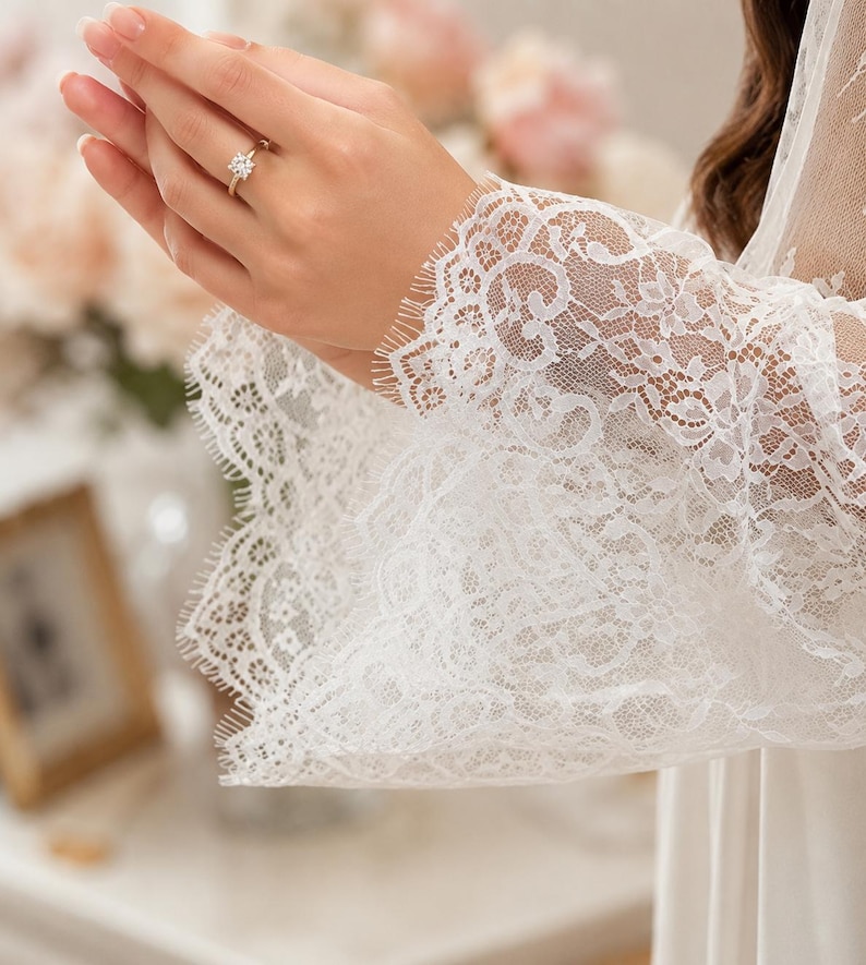 May include: A white lace robe featuring long, wide sleeves with intricate floral patterns and scalloped, fringed edges. The fabric is sheer and delicate, showcasing a detailed lace texture throughout the sleeve design and cuff area.
