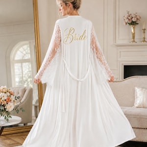 May include: A floor-length white robe featuring long, sheer lace sleeves and a matching tie belt. The back of the robe displays the word "Bride" printed in a gold, cursive script font. The garment is made of a smooth, solid white fabric.