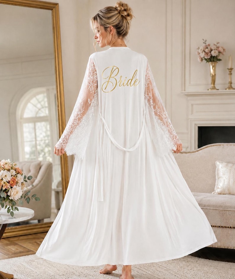 May include: A floor-length white robe featuring long, sheer lace sleeves and a matching tie belt. The back of the robe displays the word "Bride" printed in a gold, cursive script font. The garment is made of a smooth, solid white fabric.
