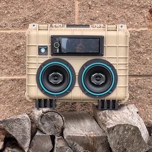 May include: A tan, rectangular, hard-shell portable speaker case featuring two circular speakers with bright blue accent rings, a central digital head unit with a black display, and a small silver power button labeled "POWER ON" on the front.