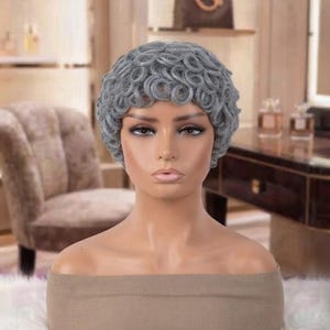 May include: A mannequin head featuring a short, curly gray wig with tight, defined ringlets. The mannequin has a smooth, light-toned complexion, neutral facial features, and is wearing a plain, beige off-the-shoulder top covering the shoulders.
