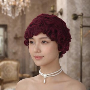 May include: A deep burgundy short wig featuring tight, textured curls styled in a rounded shape. The wig is paired with a double-strand white pearl choker necklace that includes a central decorative silver-toned clasp and a single teardrop pearl.