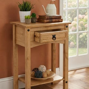 Puede incluir: Una mesa auxiliar de madera de color marrón claro con un cajón que tiene un tirador de anillo de metal negro y tachuelas decorativas en las patas. La mesa tiene un estante inferior abierto con un cuenco de madera y esferas decorativas.
