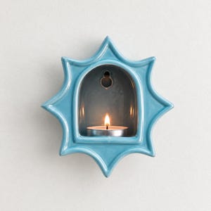 May include: A star-shaped ceramic wall sconce in a glossy light blue finish. The centre features an arched alcove containing a lit tea light candle. A small circular hole is visible at the top of the interior arch for mounting purposes.