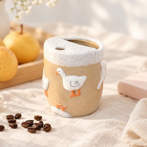 May include: A ceramic travel mug with a speckled tan body and a white speckled rim. The mug features raised, three-dimensional white goose figures with orange beaks and feet. It has a slanted top opening with a small circular hole for a straw.