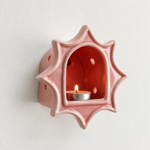 May include: A star-shaped ceramic wall sconce in a glossy dusty pink finish. The piece features a central arched opening containing a lit tea light candle and small circular ventilation holes along the sides of the structure.