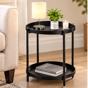 May include: A black metal two-tier round side table with a raised edge on both shelves. The top shelf holds a small lamp with a white shade, a green potted plant, and a lit candle. The bottom shelf holds two stacked books with grey covers.