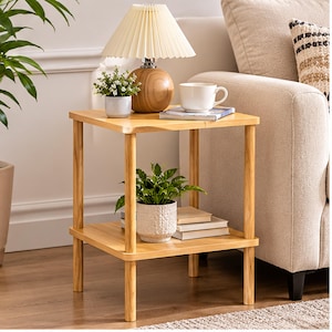May include: A square wooden side table with four cylindrical legs and a lower shelf. The top surface holds a lamp with a pleated shade, a small potted plant, and a white mug on a book. The lower shelf holds a potted plant and two books.