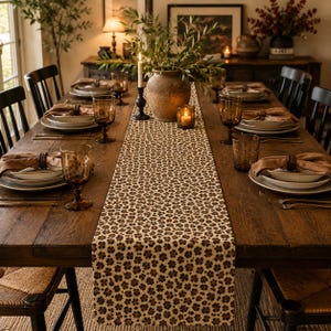 May include: A long rectangular table runner featuring a repeating pattern of small brown and black flower-like shapes on a light beige fabric. The runner is centered on a dark wooden dining table set with plates, napkins, and glassware.