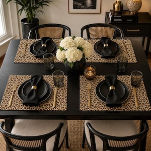 May include: A black dining table set for four with leopard print placemats, matte black plates, black napkins held by gold rings, gold forks and knives, and dark textured glasses. A black vase with white hydrangea flowers sits in the centre.