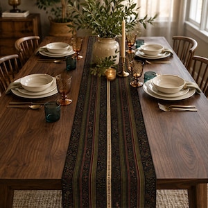 May include: A long rectangular table runner featuring a dark botanical pattern with vertical stripes in shades of black, olive green, and deep red, accented by a central cream-colored band with a delicate leaf motif, laid across a wooden table.
