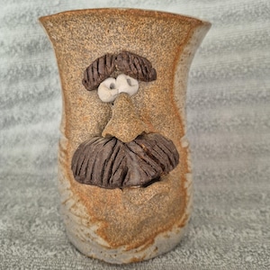 May include: A tan ceramic mug with a speckled texture featuring a sculpted face on the front. The face includes two small white eyes, a prominent nose, a dark brown textured mustache, and dark brown eyebrows, all integrated into the clay surface.
