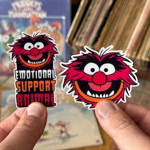 May include: Two vinyl stickers featuring the Muppets character Animal. One sticker shows the character's face with the text EMOTIONAL SUPPORT ANIMAL in white and orange block letters. The second sticker shows only the character's face with red fur.