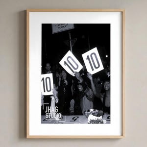 Könnte beinhalten: Ein gerahmtes Schwarz-Weiß-Foto, das eine Menschenmenge zeigt, die weiße Wertungskarten mit der in Schwarz gedruckten Zahl 10 hochhält. Der Text JHAG STUDIO DIGITAL DOWNLOAD ist in der unteren linken Ecke des Drucks in einem hellen Holzrahmen zu sehen.