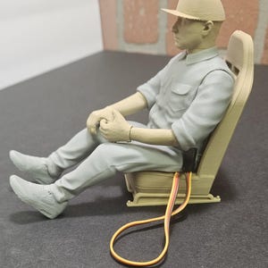 May include: A 3D-printed figurine of a person wearing a cap, collared shirt, and pants, seated in a bucket-style racing seat. The figure is light beige and gray, with a small black electronic component and attached multi-colored wires extending from the side of the seat base.