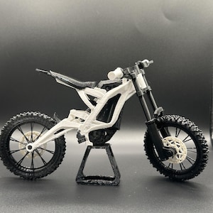 Può includere: Un modellino di moto da cross elettrica con telaio bianco, ruote nere con pneumatici tassellati, forcella anteriore nera e sella nera. Il modello è sostenuto da un cavalletto triangolare nero e presenta dettagli bianchi sui dischi freno.