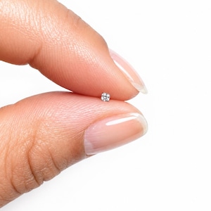 May include: A tiny, clear, round-cut gemstone stud earring featuring a single sparkling stone set in a small, polished silver-toned metal prong setting, held between two fingers against a plain white background.