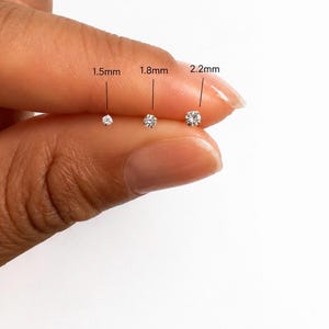 Peut inclure: Trois petites boucles d'oreilles à clous rondes et transparentes avec des montures argentées. Les clous sont étiquetés avec leurs diamètres respectifs de 1,5 mm, 1,8 mm et 2,2 mm, montrant la progression de taille de la plus petite à la plus grande.