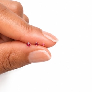 May include: Three small, round, faceted red gemstone stud earrings set in gold metal prongs. The stones are arranged in a horizontal row, decreasing in size from left to right, showcasing a vibrant crimson hue and a polished metallic finish.