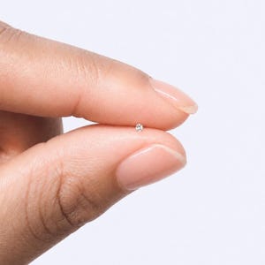 May include: A tiny, circular silver stud earring featuring a single, small, clear, faceted gemstone set in the center. The stud is held between two fingers, showcasing its miniature scale and reflective metallic finish against a plain background.