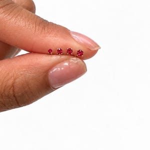 May include: Four small stud earrings featuring round, faceted red gemstones set in gold-toned metal prongs. The earrings are arranged in a horizontal row, showing a graduated size progression from the smallest on the left to the largest on the right.