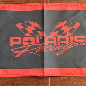 May include: A rectangular flag featuring a black center with the red text Polaris Racing and two red and black checkered flags above it. The flag has red horizontal borders at the top and bottom, and a black vertical strip on the left with three brass grommets.