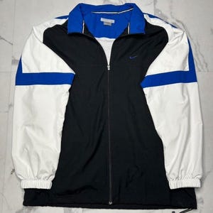 May include: A full-zip windbreaker jacket featuring a color-blocked design in black, white, and royal blue. The jacket has a high collar, long sleeves with elastic cuffs, and a small embroidered Nike swoosh logo on the upper left chest area.
