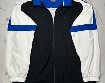 May include: A full-zip windbreaker jacket featuring a color-blocked design in black, white, and royal blue. The jacket has a high collar, long sleeves with elastic cuffs, and a small embroidered Nike swoosh logo on the upper left chest area.