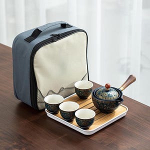 May include: A tea set featuring a dark blue ceramic teapot with a wooden side handle and a small red knob on the lid, accompanied by four matching cups. The set sits on a rectangular bamboo tray and includes a grey and beige fabric carrying case.