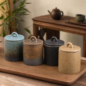 May include: Four cylindrical ceramic canisters with lids, each featuring a small arched handle. The canisters display varied textures and colors, including blue-to-black gradient, brown-to-gray gradient, solid matte black, and a speckled tan finish.