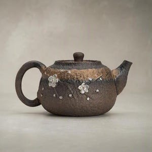 May include: A dark brown, textured ceramic teapot featuring a rounded body, a curved handle, and a short spout. The surface is decorated with small, raised silver-coloured floral accents and thin branches that wrap around the exterior of the pot.