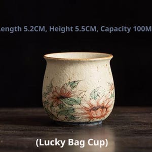 May include: A cream-colored ceramic cup with a textured surface and a floral pattern featuring orange and green tones. The cup is labeled Lucky Bag Cup and measures 5.2 cm in length, 5.5 cm in height, and has a capacity of 100 ml.