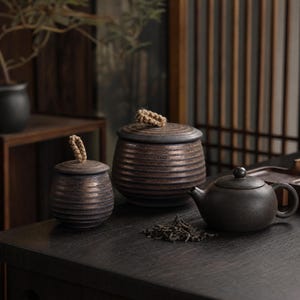 May include: Two dark brown ceramic canisters with horizontal ribbed textures and small rope handles on their lids, accompanied by a smooth dark brown ceramic teapot and a small pile of loose tea leaves on a dark wooden surface.