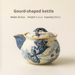 Peut inclure: Une bouilloire en forme de gourde avec une base crème, des motifs de papillons et de nuages bleus, et une finition mouchetée. Le texte indique Gourd-shaped kettle, Width 10.7cm, Height 6.5cm, Capacity 200ml. Elle possède un couvercle et un bec bleu.