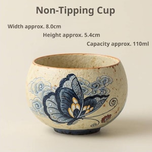 May include: A beige ceramic non-tipping cup with a speckled texture, featuring a blue and yellow butterfly graphic and stylized clouds. Text reads: Non-Tipping Cup, Width approx. 8.0cm, Height approx. 5.4cm, Capacity approx. 110ml. The cup has a rounded shape.
