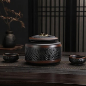 Puede incluir: Un bote de té de cerámica en color marrón oscuro y negro con un patrón de tejido texturizado en el cuerpo y la tapa, que presenta un pequeño asa de anillo de latón en la parte superior, acompañado de dos cuencos de cerámica a juego.