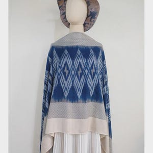 May include: A blue and beige patterned shawl draped over a mannequin wearing a matching wide-brimmed hat and a white pleated skirt. The shawl features a prominent diamond-shaped ikat-style design in shades of blue, transitioning into solid beige bands at the bottom hem.