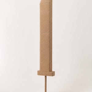 Puede incluir: Una espada grande construida con cartón de color marrón claro. La hoja está compuesta por varios segmentos rectangulares apilados verticalmente, con un borde superior plano y una guarda rectangular. Un mango cilíndrico se extiende hacia abajo.