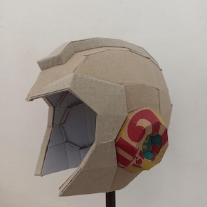 May include: A geometric helmet constructed from light brown cardboard panels. The helmet features a prominent visor section and a circular side component decorated with red, yellow, and blue printed patterns. The interior structure is visible.