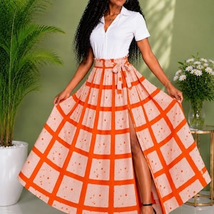 May include: A white short-sleeved button-down shirt paired with a high-waisted, floor-length maxi skirt featuring a bold orange grid pattern on a light beige background, a tie-waist detail, and a high front slit that reveals the leg.