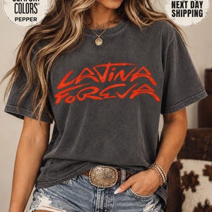 May include: A charcoal gray short-sleeve t-shirt featuring the text "LATINA FOREVA" in bold red lettering across the chest. The shirt has a crew neckline and a relaxed fit, paired with distressed blue denim shorts and a large ornate metal belt buckle.