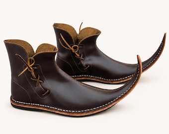 May include: A pair of dark brown leather ankle boots featuring an elongated, pointed toe design that curves upward. The boots have a side lace-up closure with thin leather cords and visible white stitching along the sole edge and base.