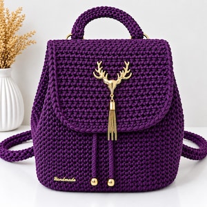 Peut inclure: Un sac à dos en crochet violet foncé orné d'un emblème en métal doré en forme de bois de cerf avec une pampille à chaînette. Le sac possède une poignée, deux bretelles, une fermeture à cordon avec des perles dorées et une étiquette "Handmade".