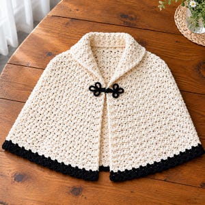 May include: A cream-colored crochet capelet featuring a textured stitch pattern, a wide collar, and a black decorative frog closure at the neckline. The hem of the garment is finished with a solid black crochet border that contrasts with the cream body.