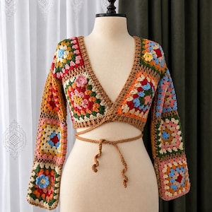 May include: A long-sleeved wrap top made of multicolored crochet granny squares. The garment features a deep V-neckline, long sleeves, and a brown yarn tie closure at the waist. The squares incorporate various colors including red, blue, yellow, pink, and green.