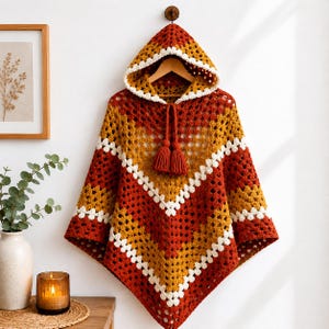 May include: A hooded crochet poncho featuring a chevron pattern in alternating stripes of rust orange, mustard yellow, and white. The garment includes a drawstring hood with two matching rust orange tassels hanging from the center neckline.