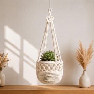 Peut inclure: Une suspension pour plante en macramé couleur crème avec un nœud central en forme de fleur au sommet, des motifs en treillis tissés complexes au milieu et une base solide, contenant une succulente verte dans un pot blanc.