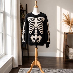 Könnte beinhalten: Ein schwarzer Langarmpullover mit einem markanten weißen Skelett-Design aus Brustkorb und Beckenknochen auf der Vorderseite. Die Ärmel zeigen weiße Armknochen-Grafiken. Das Kleidungsstück wird auf einer hölzernen Schaufensterpuppe präsentiert.