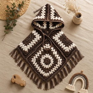 Op de afbeelding: Een poncho met capuchon gemaakt van donkerbruin en wit garen in een granny square haakpatroon. Het kledingstuk heeft een puntige zoom afgewerkt met lange bruine franjes en is voorzien van een bijpassend trekkoord bij de halslijn.